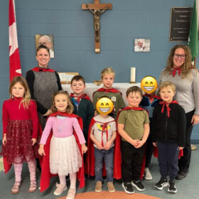 Groupe d’élèves portant des capes rouges posant avec deux adultes dans une salle décorée avec un drapeau canadien et un crucifix.