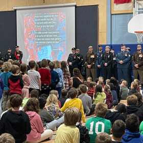 Élèves réunis dans un gymnase devant un groupe de militaires lors d’une cérémonie du jour du Souvenir, avec un grand écran affichant un poème et des coquelicots.